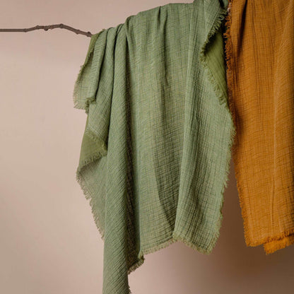 Two sage green linen throws with hand-pulled fringe details hanging side by side on wooden hooks, highlighting their artisanal texture.